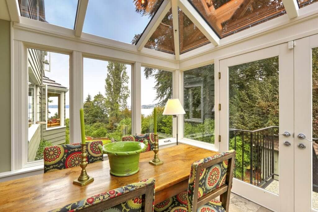 Sunroom patio area with transparent vaulted ceiling, wooden dining table with colorful chairs. Exit to backyard