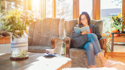 Cheerful smiling female dressed cozy home clothes sitting in comfortable armchair, relaxing, reading a book and drinking morning coffee or tea in house sunroom living room. Stay-at-home concept image